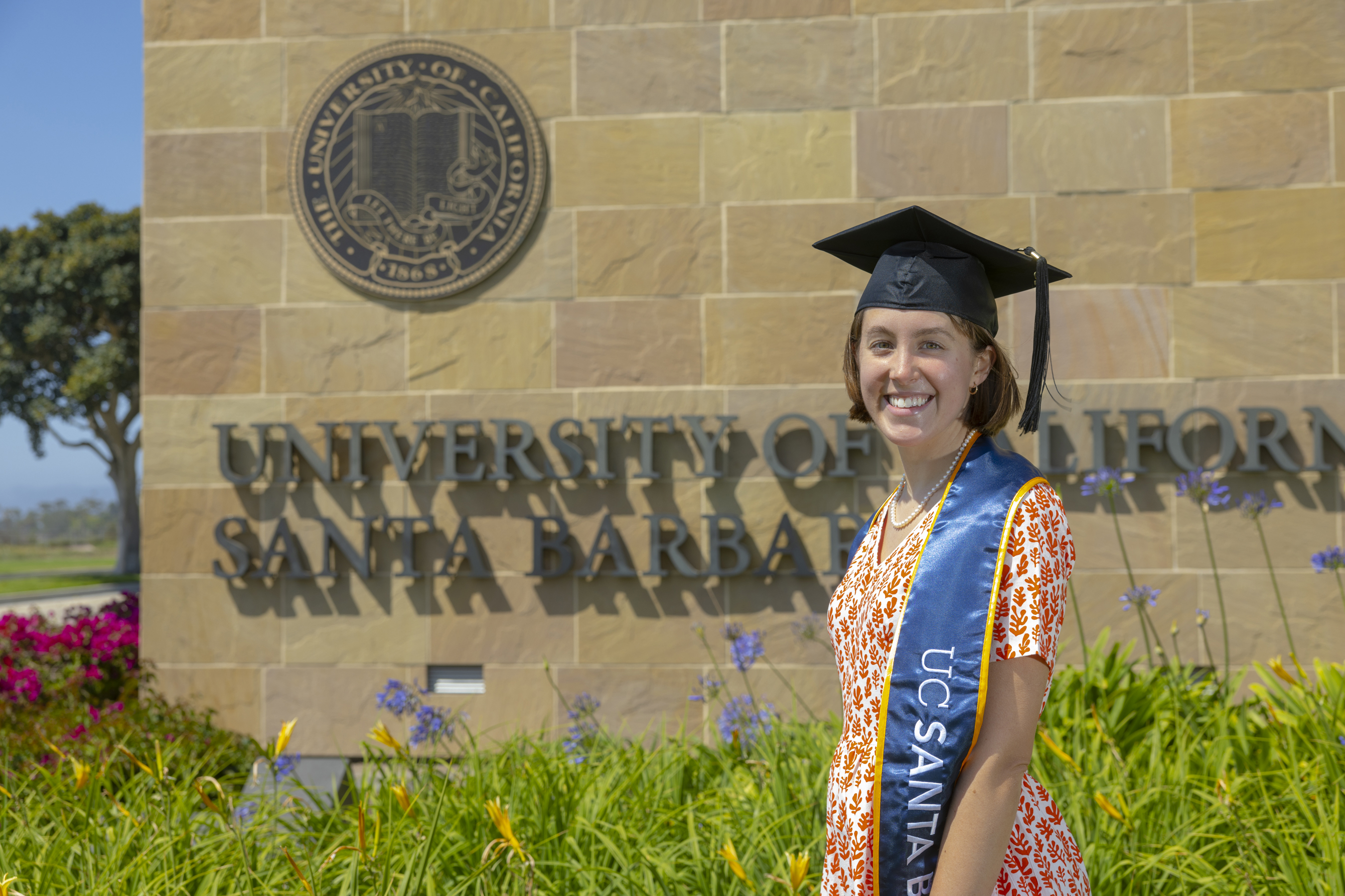 Scout dressed in graduation regalia for graduating from her Master degree. Scout wears a black grad cap and a red Matrisse-esque print dress. She stands in front of a sand-colored stone wall that reads 'University of California Santa Barbara,' framed by blue skies and green grass.