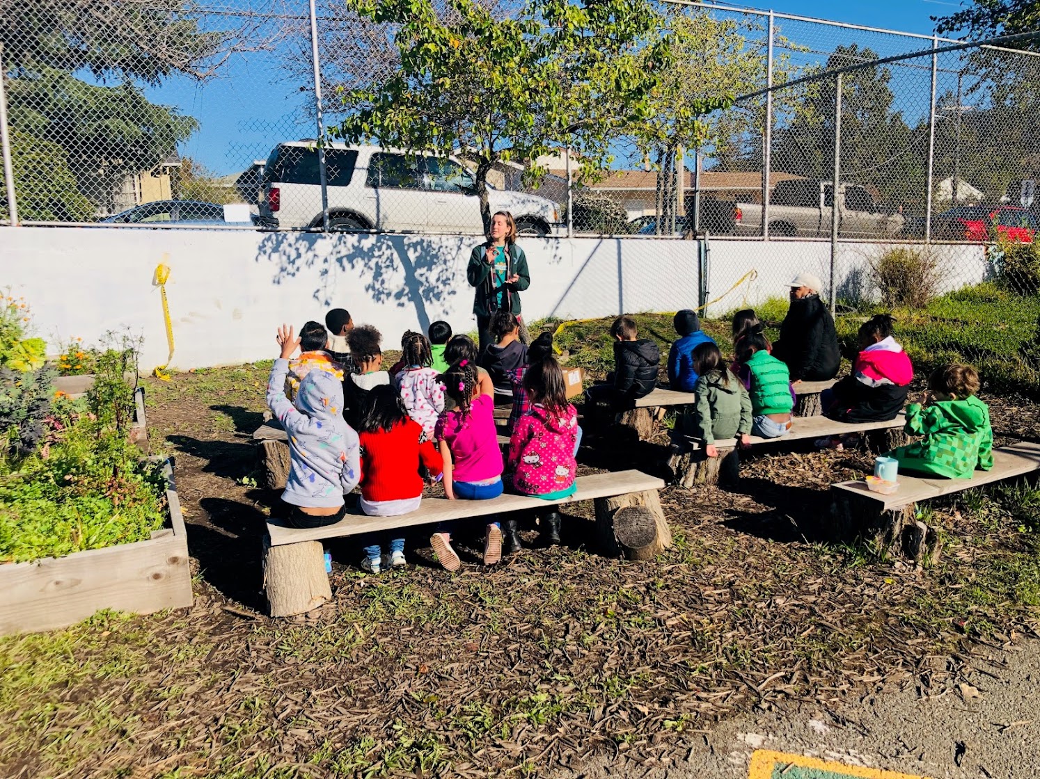 Prior to managing and implementing district-wide school meal programs in Oakland, Scout worked closely with students in school gardens and cafeterias! In this photo she stands in front of a class of 5-6 year-olds who are in a school garden of an Oakland neighborhood.